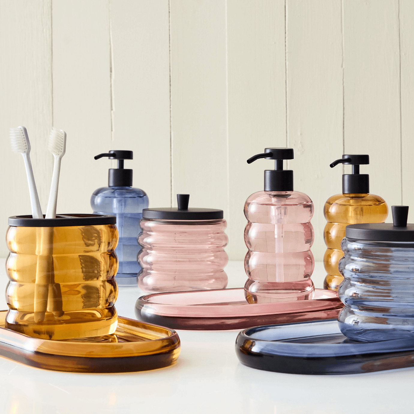 12 Colored Glass Accessories We Love: Cake Stands, Candlesticks & More - 5 Assorted colored glass items, including ribbed soap dispensers, tumblers with toothbrushes, and trays in amber, pink, and blue hues, make a chic addition to any spring wish list. These vibrant bathroom accessories shine against a light wooden background.