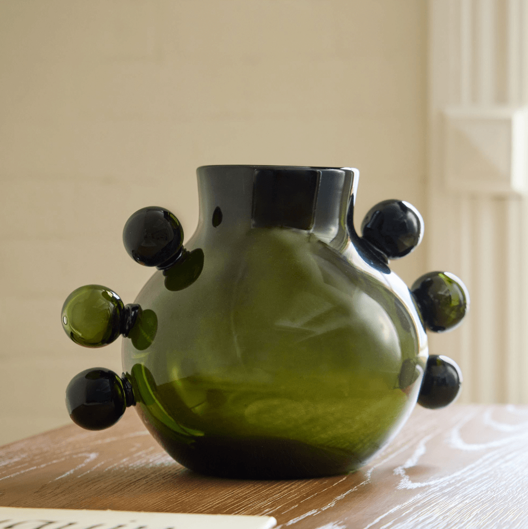 12 Colored Glass Accessories We Love: Cake Stands, Candlesticks & More - 1 A green glass vase with bulbous protrusions sits on a wooden table indoors, adding a touch of fresh elegance.