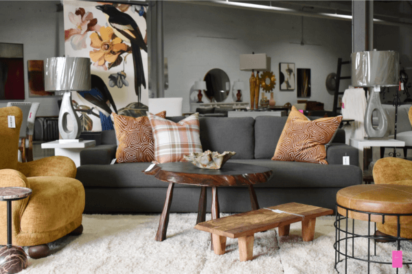 A modern living room showroom by So So Spiffy, featuring a grey sofa, patterned pillows, a wooden coffee table, mustard armchairs, and decorative lamps on a white rug—perfect inspiration for your next shopping shindig.