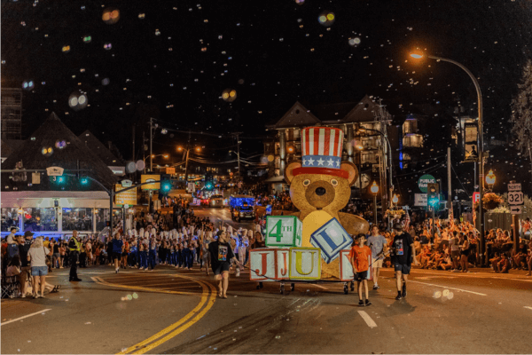 People in Gatlinburg march at night behind a large teddy bear float in a patriotic hat during the 4th of July Parade, with crowds lining the street and bubbles floating through the air.