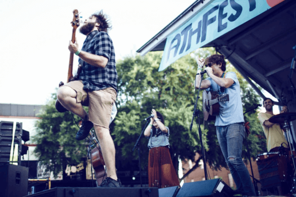 A band performs energetically on an outdoor stage at the AthFest Music Festival, with musicians playing guitar and bass beneath a banner reading "ATHFEST" overhead.