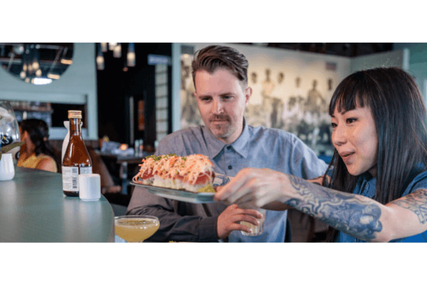 A woman serves a plate of food to a man at a restaurant bar. Both are seated with drinks, enjoying the casual atmosphere and special restaurant deals during Athens Restaurant Week.