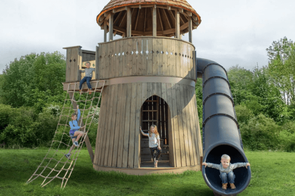 Children play on a wooden enchanted castle-themed playground at Cheekwood, with one boy on a slide, two kids on a rope ladder, and a girl in the tower entrance; all surrounded by grass and trees.