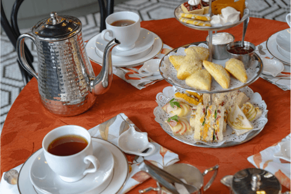 A table set for afternoon tea at Cheekwood features a silver teapot, two cups of Summer Afternoon Iced Teas, and a tiered tray holding assorted sandwiches, pastries, and scones.