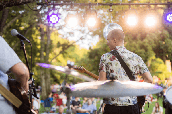 A band performs on an outdoor stage in daylight at Thursday Night Out, with bright stage lights and an audience seated on the grass in the background.