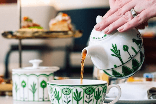 A hand pours tea from a white teapot with green patterns into a matching teacup; pastries sit on a stand in the background, capturing the elegance of a Bridgerton Afternoon Tea.