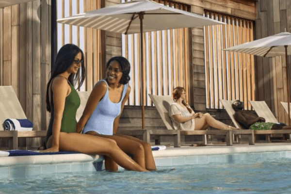 Two women sit at the edge of a pool with their feet in the water, enjoying a relaxing Memorial Day pool party as two others lounge under umbrellas in the background.