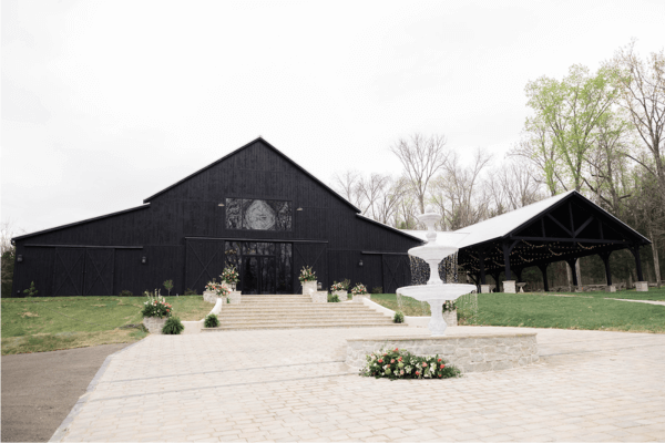 A large black barn-style building at Saddle Woods Farm, featuring glass doors, a tiered white fountain, and floral arrangements on a paved courtyard, surrounded by trees and a covered patio structure—the perfect setting for your Grand Opening.