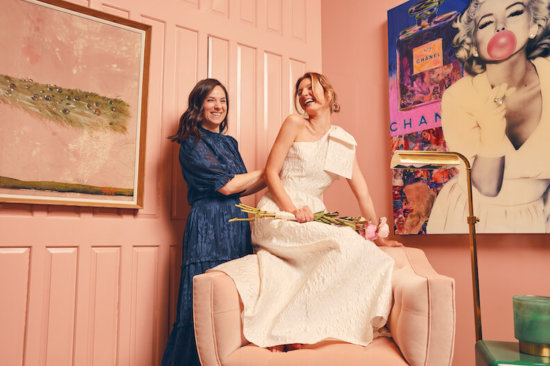 Two women, one in a blue dress and one in white, holding flowers, smile together in a stylish pink room with artwork and a floor lamp.