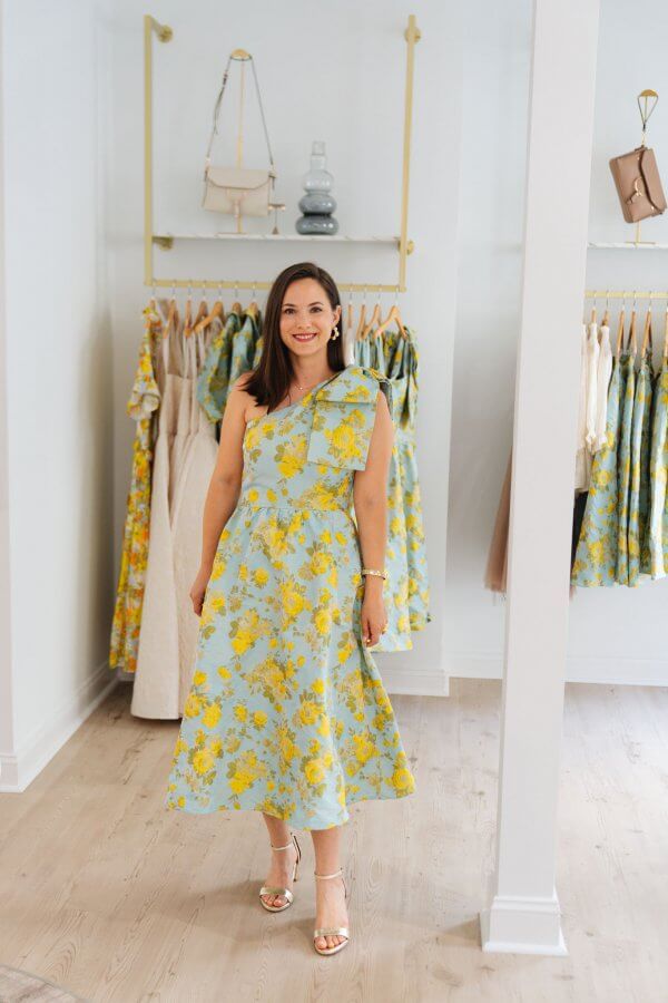 A woman stands indoors wearing a light blue, one-shoulder, yellow floral dress and gold heels, with similar dresses hanging on racks behind her.