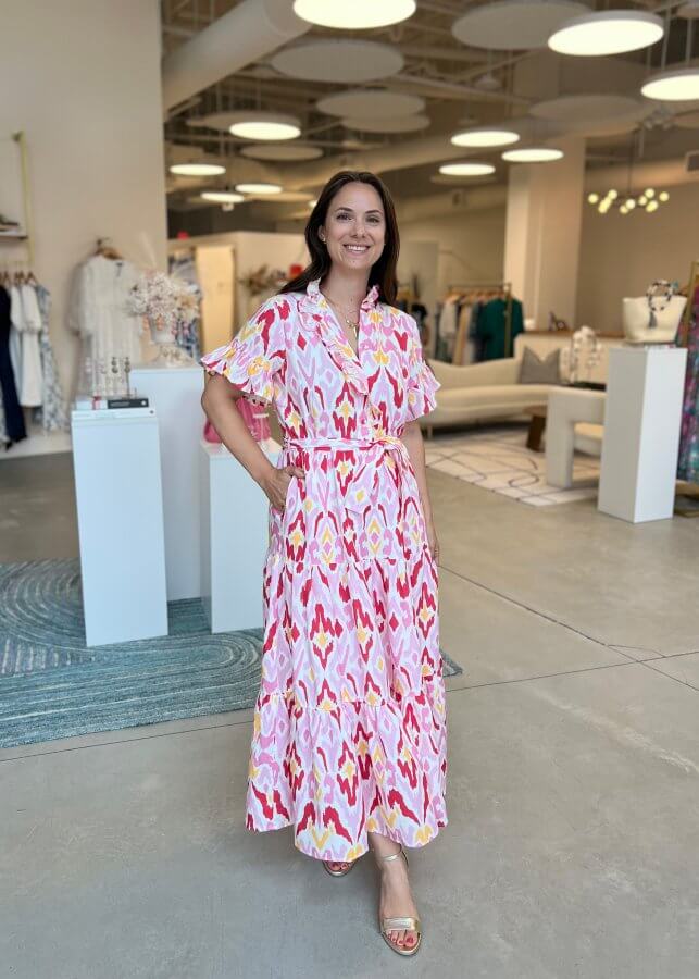A woman stands in a boutique wearing a maxi dress with a pink and yellow pattern. She is smiling, one hand on her hip, with stylish clothing displays visible in the background.