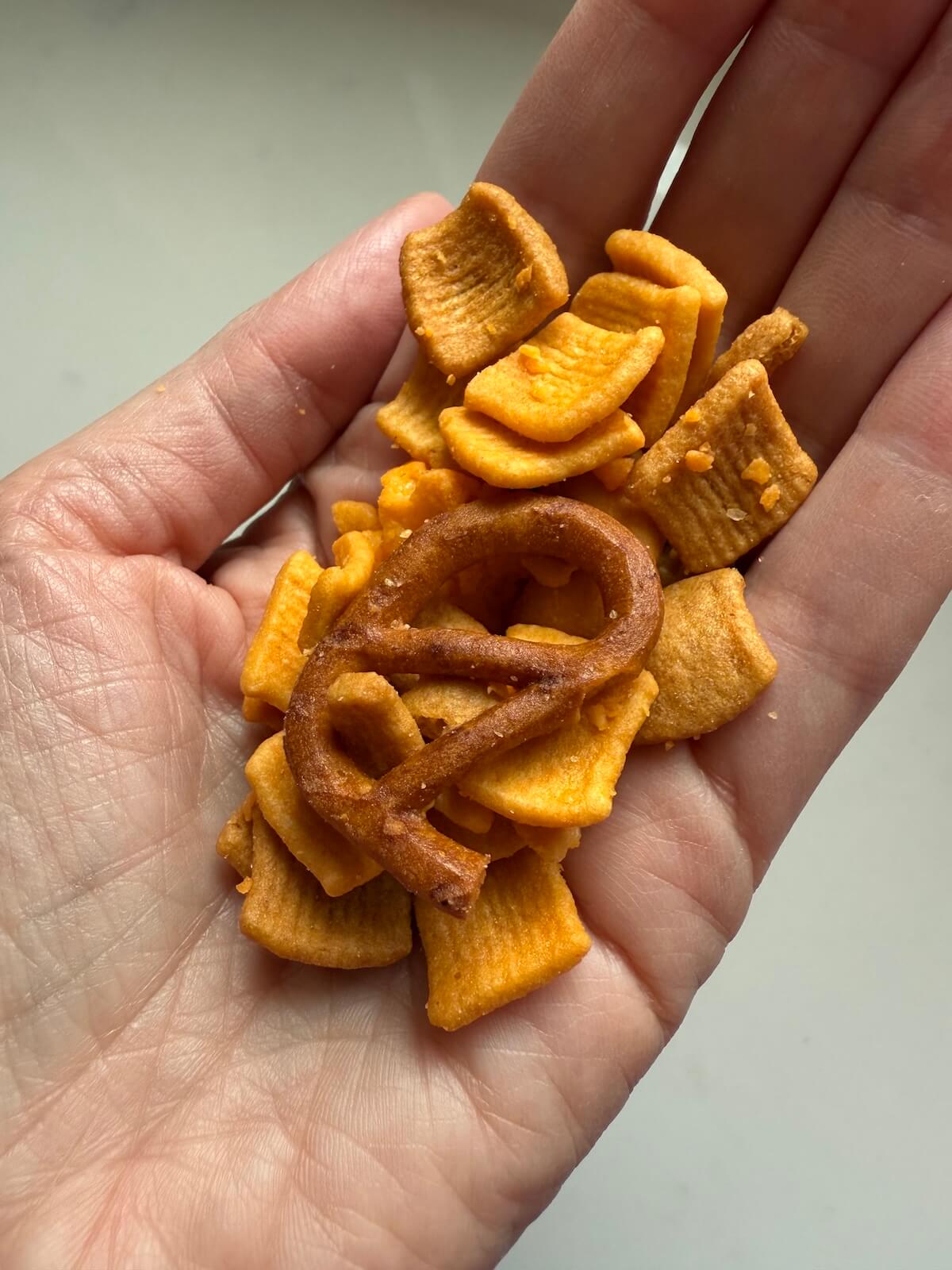 A hand holding a snack mix with square-shaped crackers and a single mini pretzel.