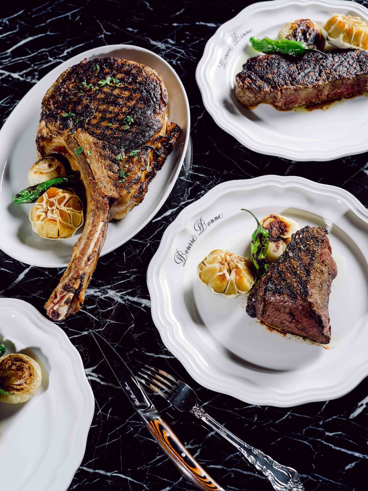 Three plates of grilled steaks with charred marks, served with roasted garlic and green pepper, on a black marble table—a tempting dish you might find at new Louisville restaurants. A fork and knife rest nearby, ready for your first bite.