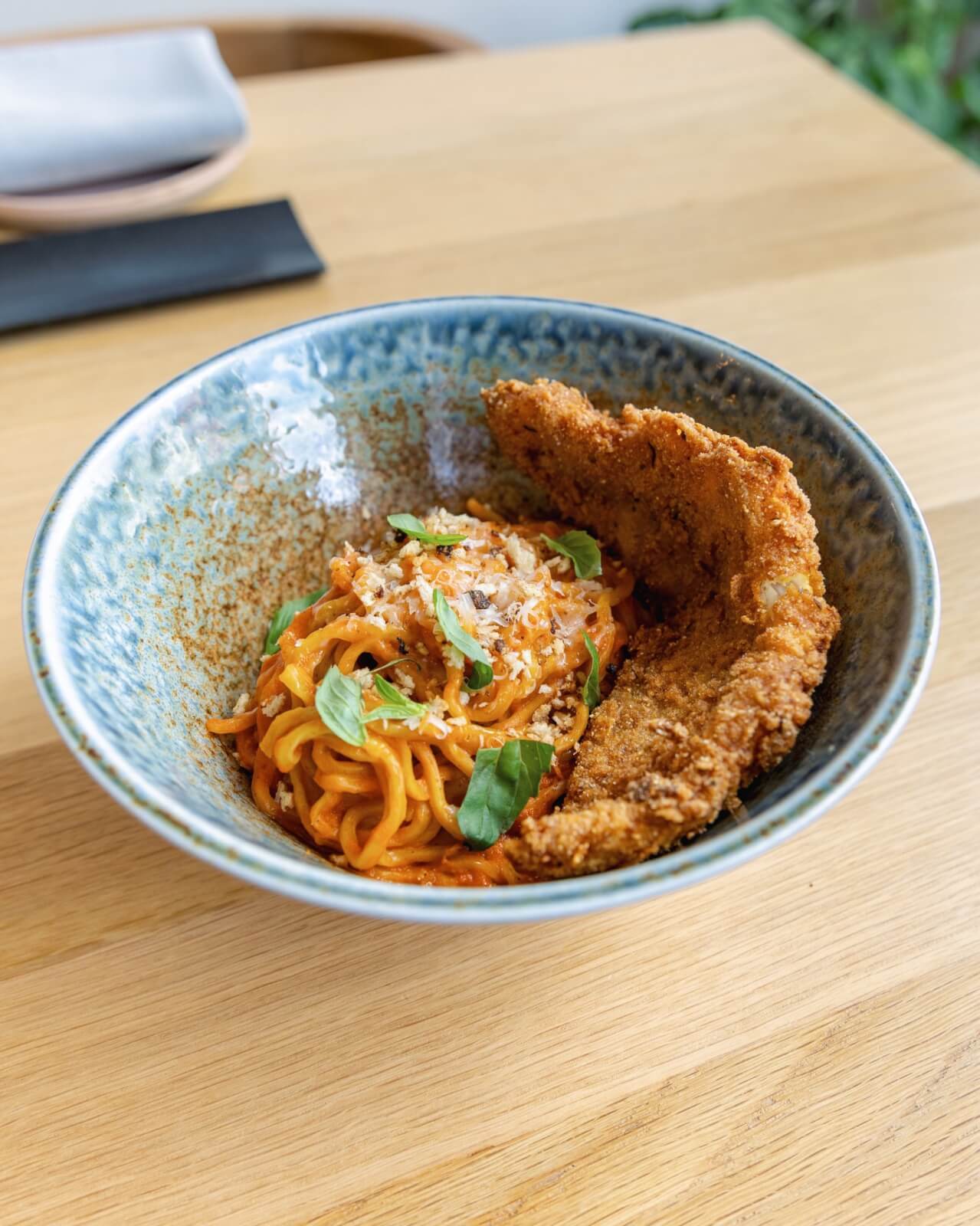 A bowl of spaghetti in tomato sauce is topped with grated cheese, basil leaves, and a breaded, fried fish cutlet.