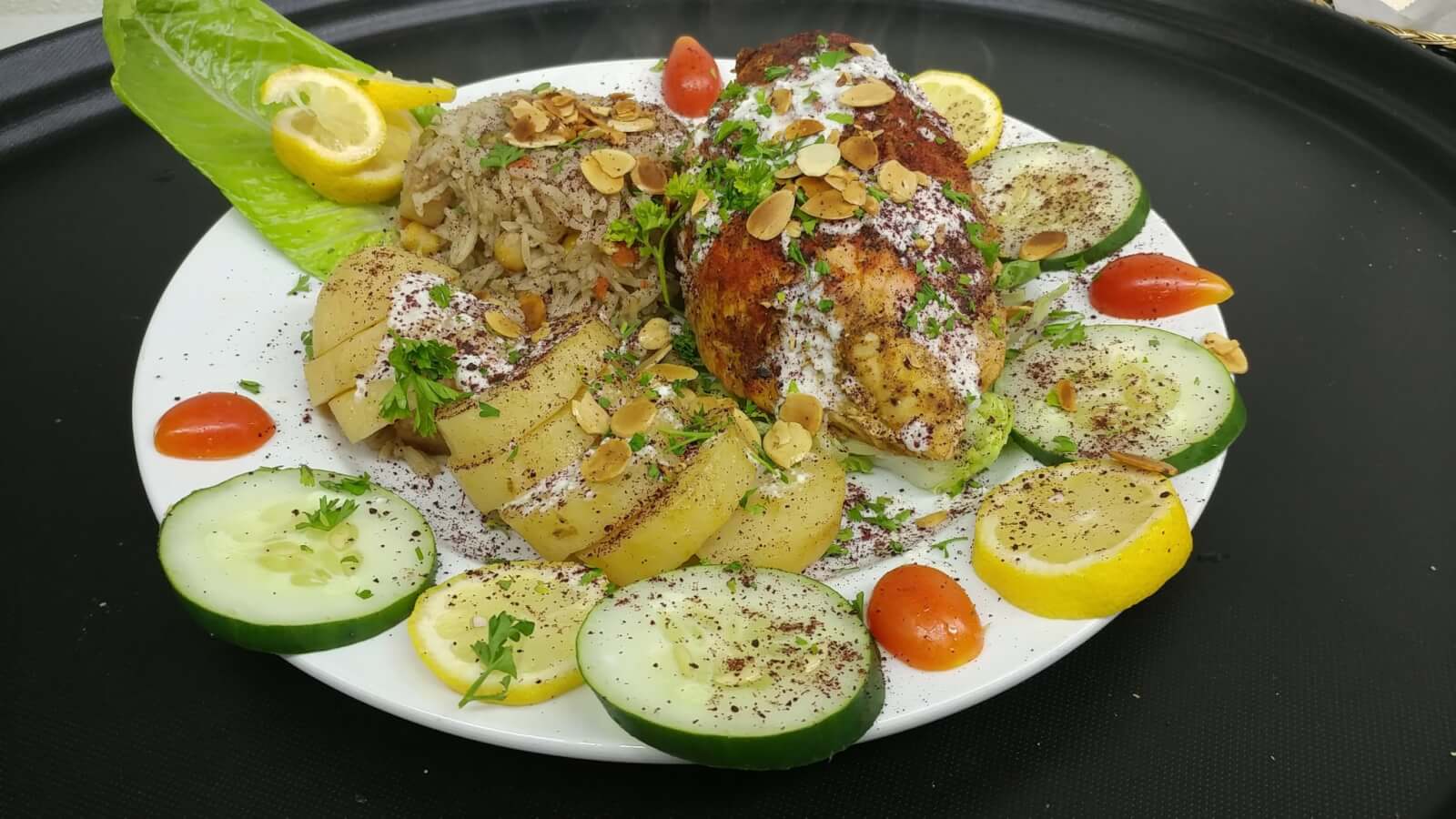 A plated meal features seasoned chicken breast with sauce, rice with almonds, sliced potatoes, cucumber, cherry tomatoes, and lemon slices, all garnished with herbs.