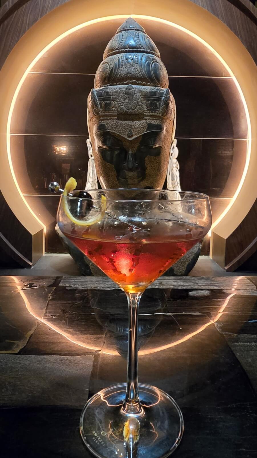 A cocktail garnished with a lemon twist and cherry sits on a reflective surface in front of a large Buddha statue with a circular light backdrop, capturing the vibrant spirit of new Louisville restaurants.