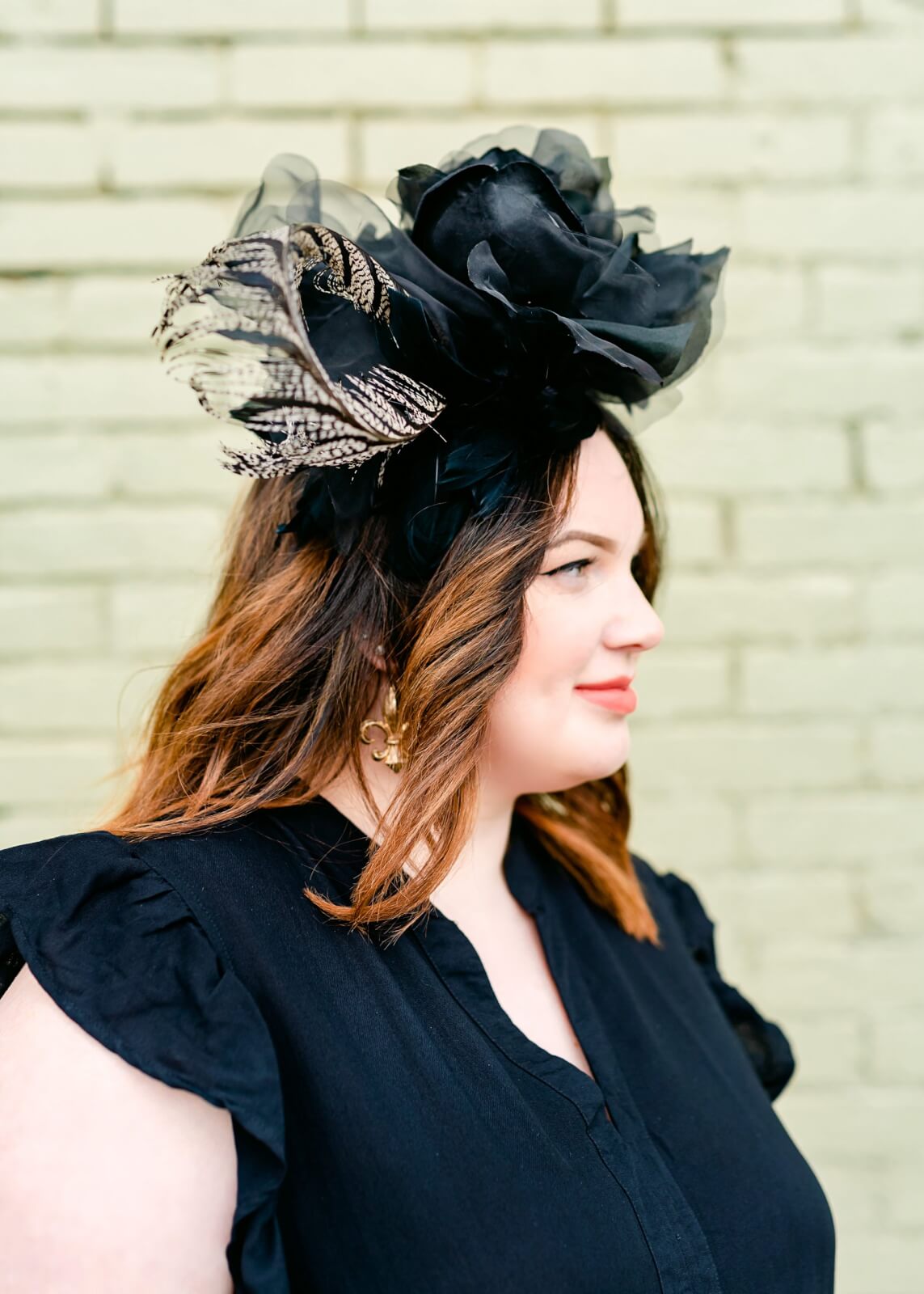 A person with medium-length hair, styled with care, is wearing a large black hat adorned with feathers and flowers. They don a sleek black top while posing against the light green brick wall of Louisville’s TRIM NuLu district, capturing a unique fusion of style and setting.