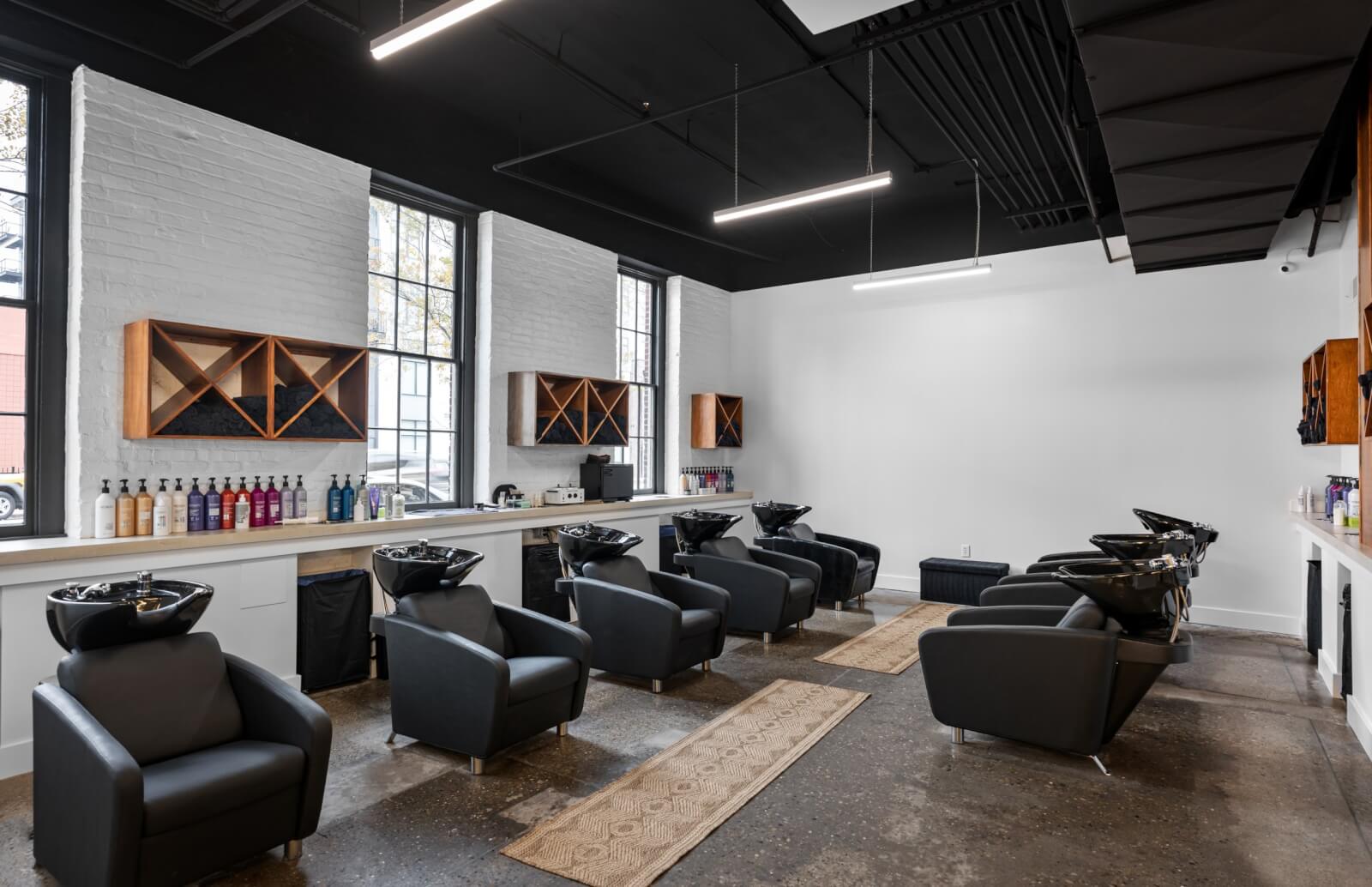 The modern salon interior of TRIM NuLu in Louisville boasts six sleek black washing stations and displays an array of hair products on wooden shelves. Large windows flood the space with light, highlighting the polished concrete floors and stylish overhead lighting.