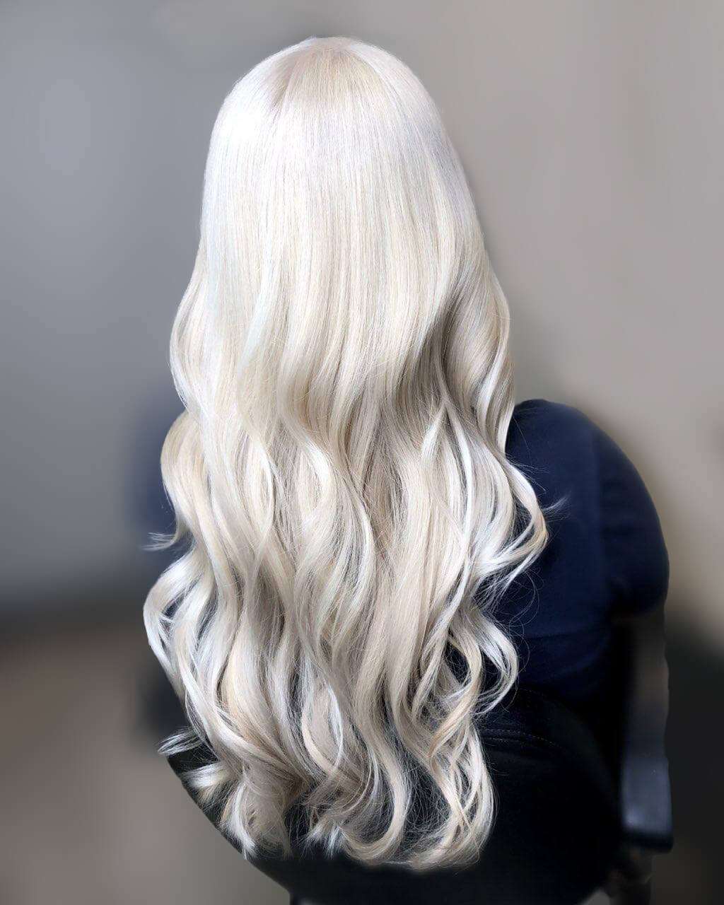 A person with long, wavy platinum blonde hair sits gracefully against a blurred gray backdrop, resembling a TRIM NuLu trend straight out of Louisville.