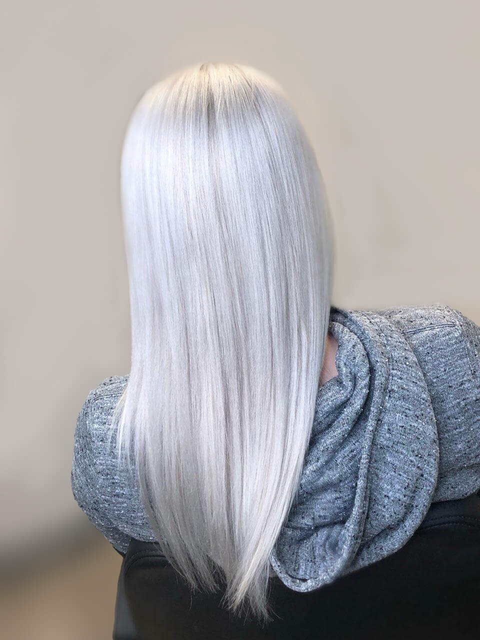 A person flaunts straight, long platinum blonde hair, perfectly styled and subtly trimmed at TRIM NuLu in Louisville, while wearing a gray hooded garment. The view from behind captures the elegance and precision of their fresh look.