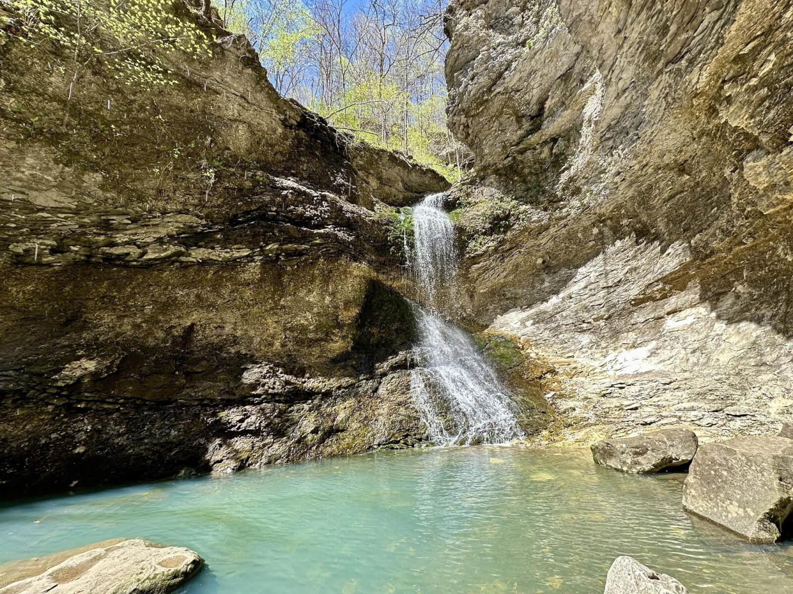 A small waterfall flows into a turquoise pool, surrounded by rocky cliffs and sparse greenery under a clear blue Arkansas sky, offering an idyllic spot for waterfall hikes.
