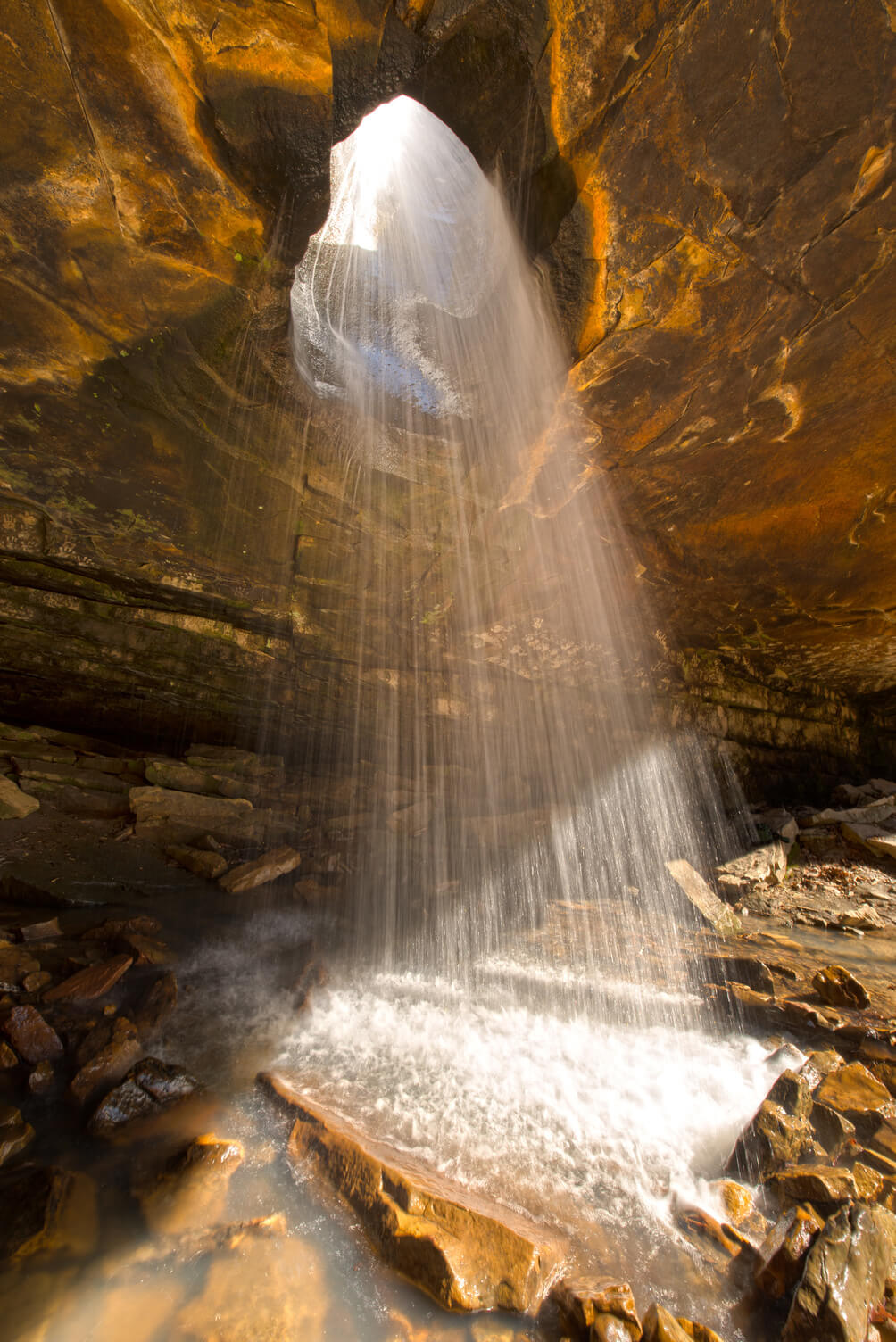Water cascades through a large hole in the rocky ceiling, creating a stunning waterfall onto the stones below. Sunlight filters through, highlighting the golden tones of the rocks—a perfect scene for those who enjoy Arkansas waterfall hikes.