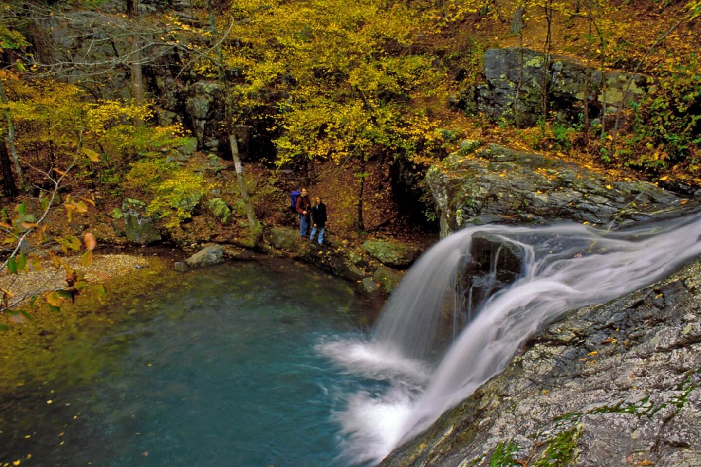 In Arkansas, a small waterfall hikes its way into a clear pool, surrounded by rocks and trees adorned with autumn leaves. Two people stand near the water's edge, soaking in nature’s tranquil beauty.