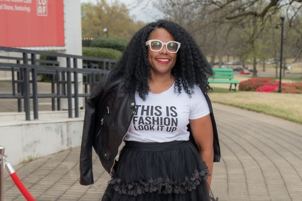 Rachel Knox, with her curly hair and glasses, confidently stands on the paved path, sporting a white shirt that declares "This Is Fashion Look It Up." Her black leather jacket and skirt complete the ensemble, adding a bold touch to her look.