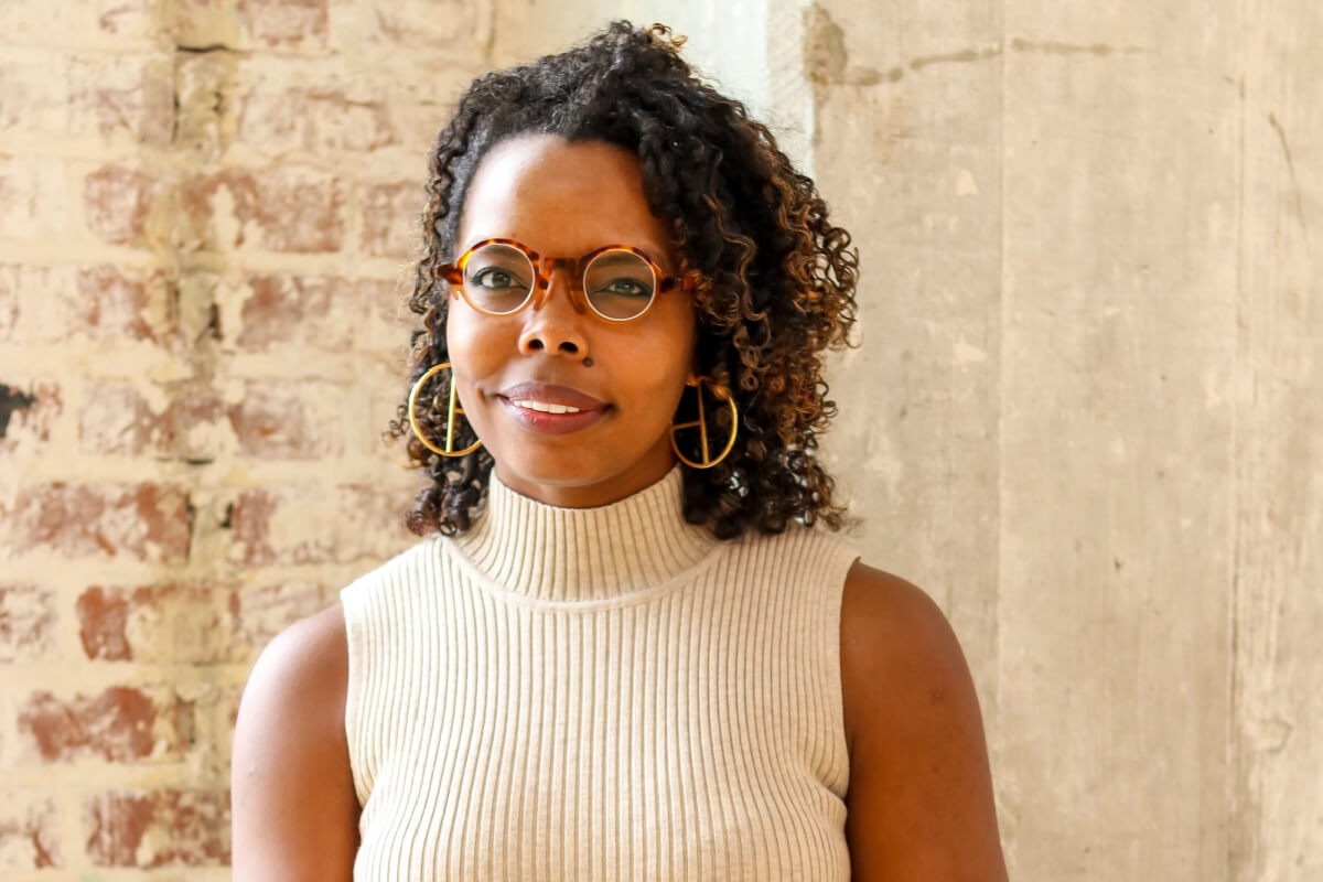 Audrey Willis, a woman with curly hair, wearing round glasses, large hoop earrings, and a sleeveless beige turtleneck, stands in front of a brick and concrete wall.