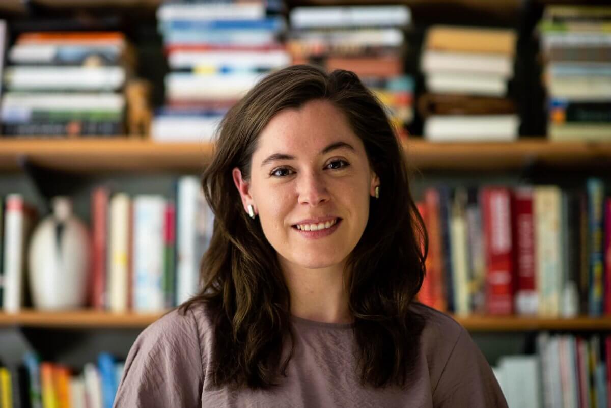 Martha Park, with long brown hair, wearing a mauve shirt and wireless earbuds, stands in front of a bookshelf filled with books—a serene portrait among the many FACES that fill the room.