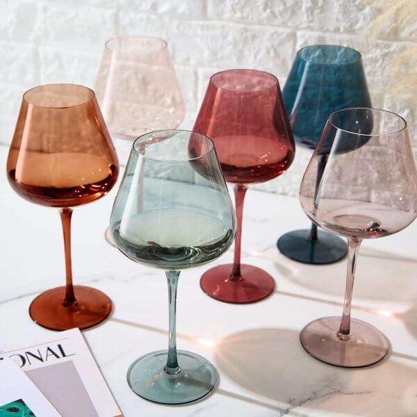 Six colored wine glasses in shades of red, pink, teal, and blue are arranged on a white marble surface near magazines and a white brick wall.
