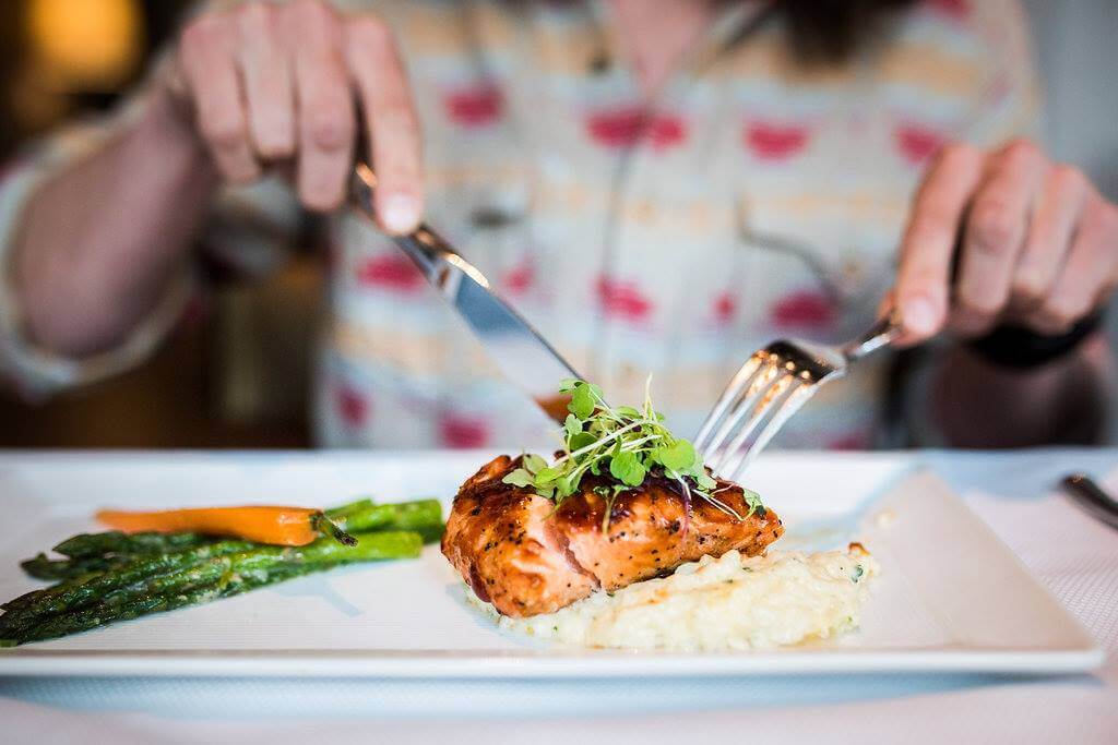 A person cuts into a piece of grilled salmon served with risotto, asparagus, and a carrot on a rectangular white plate at a cozy Memphis Mother's Day Brunch.