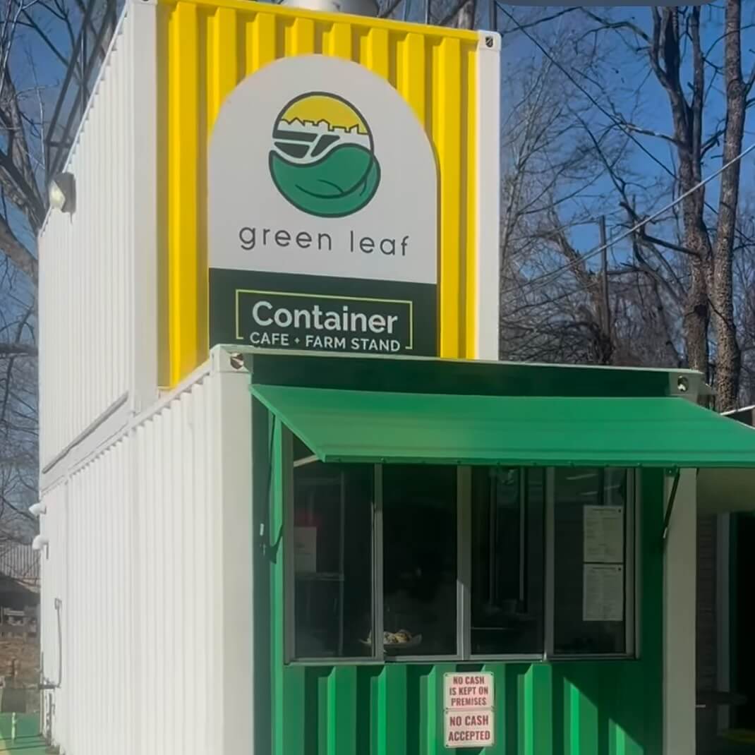 A white shipping container cafe with a green awning and a "Green Leaf" sign has joined the scene of new Memphis restaurants this April. A small window serves eager customers, while trees provide a lush backdrop.