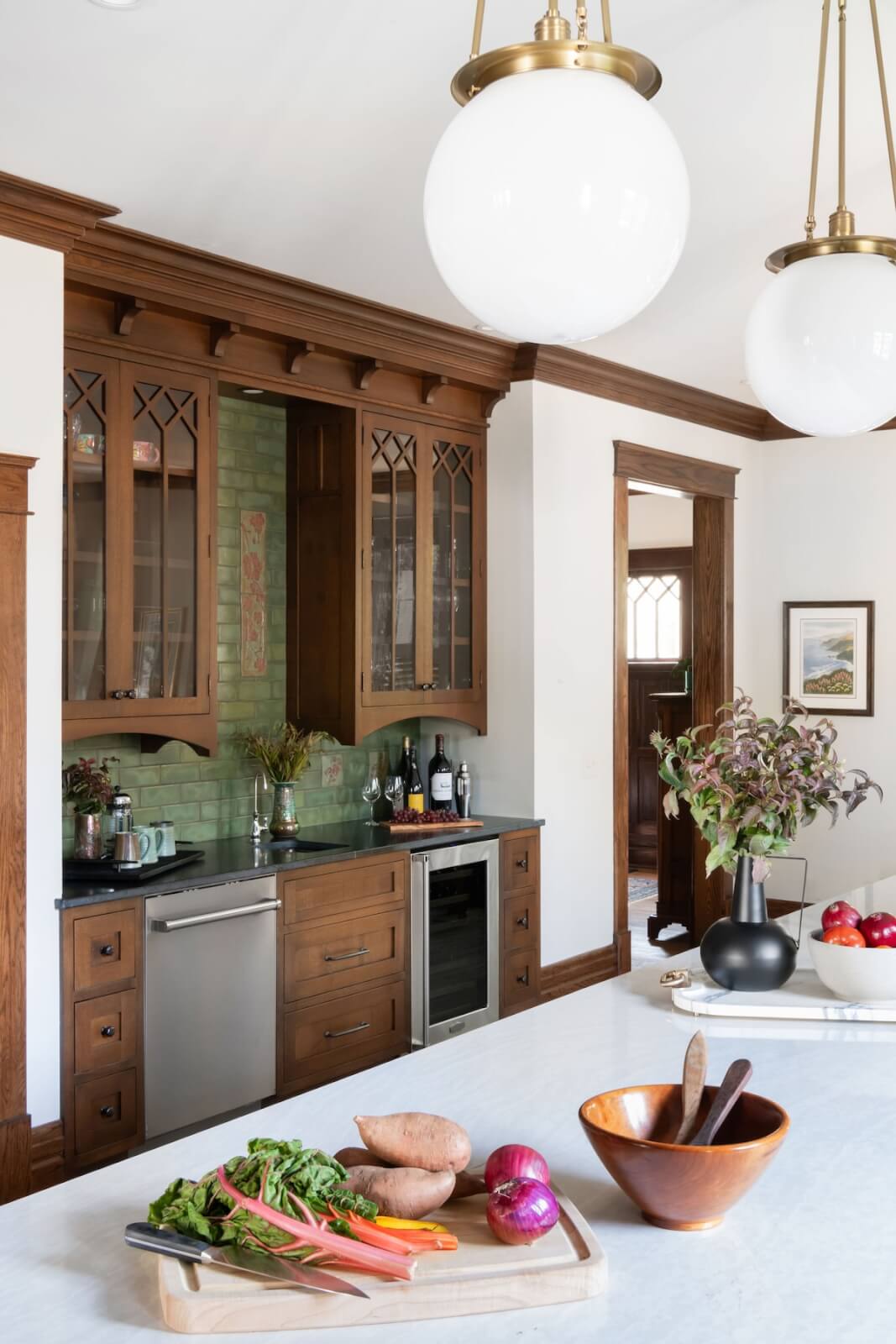 2 Homes by Beth Haley Design: A Study in Style & Contrast - 11 The kitchen interior, inspired by Beth Haley Design, features a wooden cabinet, wine bottles, and a countertop adorned with fresh vegetables on a cutting board. Decorative plants add charm as hanging globe lights beautifully illuminate the space.