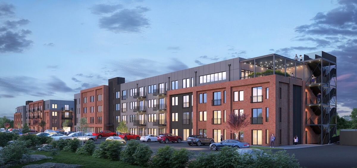 A modern four-story apartment building with brick and panel exterior, a parking lot with cars, and people on a rooftop patio at dusk.