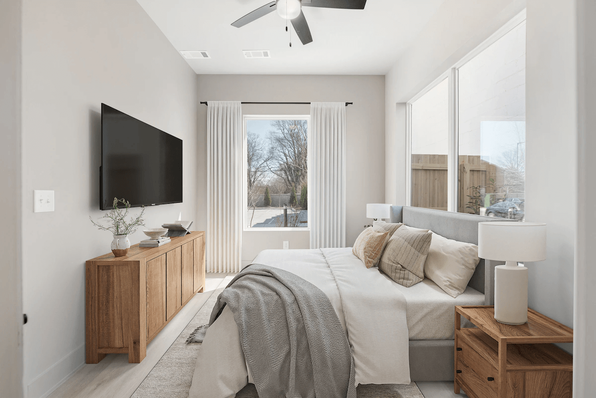 Modern East Nashville bedroom with a large bed, wooden furniture, a wall-mounted TV, neutral decor, and large windows with white curtains letting in natural light—perfect for homes in new developments.