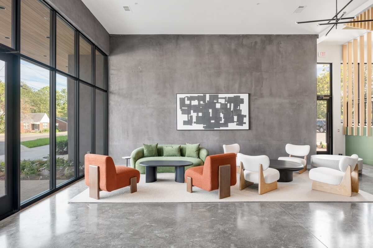 Modern lounge area in East Nashville features two orange chairs, three white chairs, a green sofa, round tables, neutral rug, and abstract artwork on a gray wall beside large windows—perfect for relaxing in new developments or stylish homes.