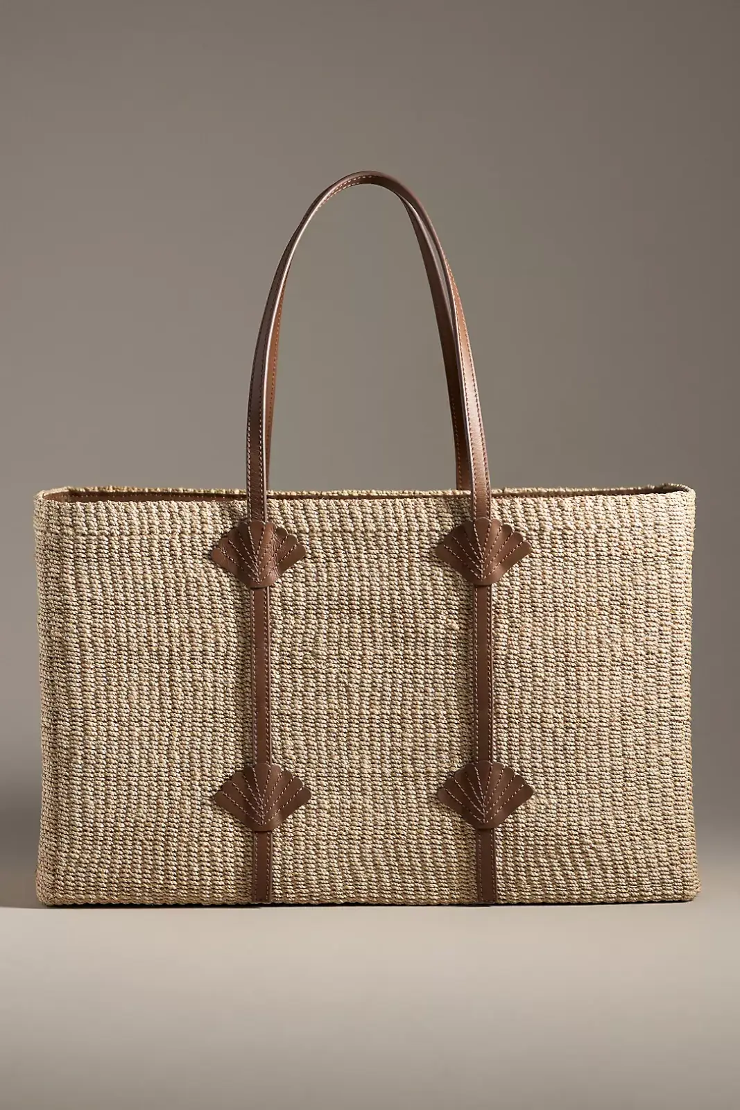 Rectangular woven straw tote bag with two brown leather handles and leather corner accents—an essential accessory for stylish summer outfits, displayed against a neutral background.