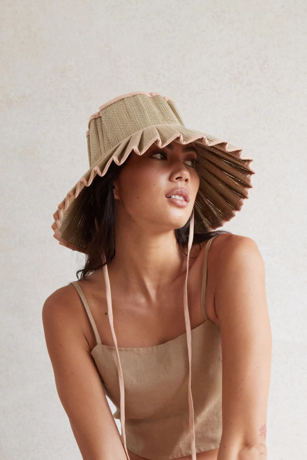 A woman wearing a light brown spaghetti strap top and a wide-brimmed, ruffled straw hat with chin ties showcases stylish summer fashion, looking slightly to the side against a neutral background.