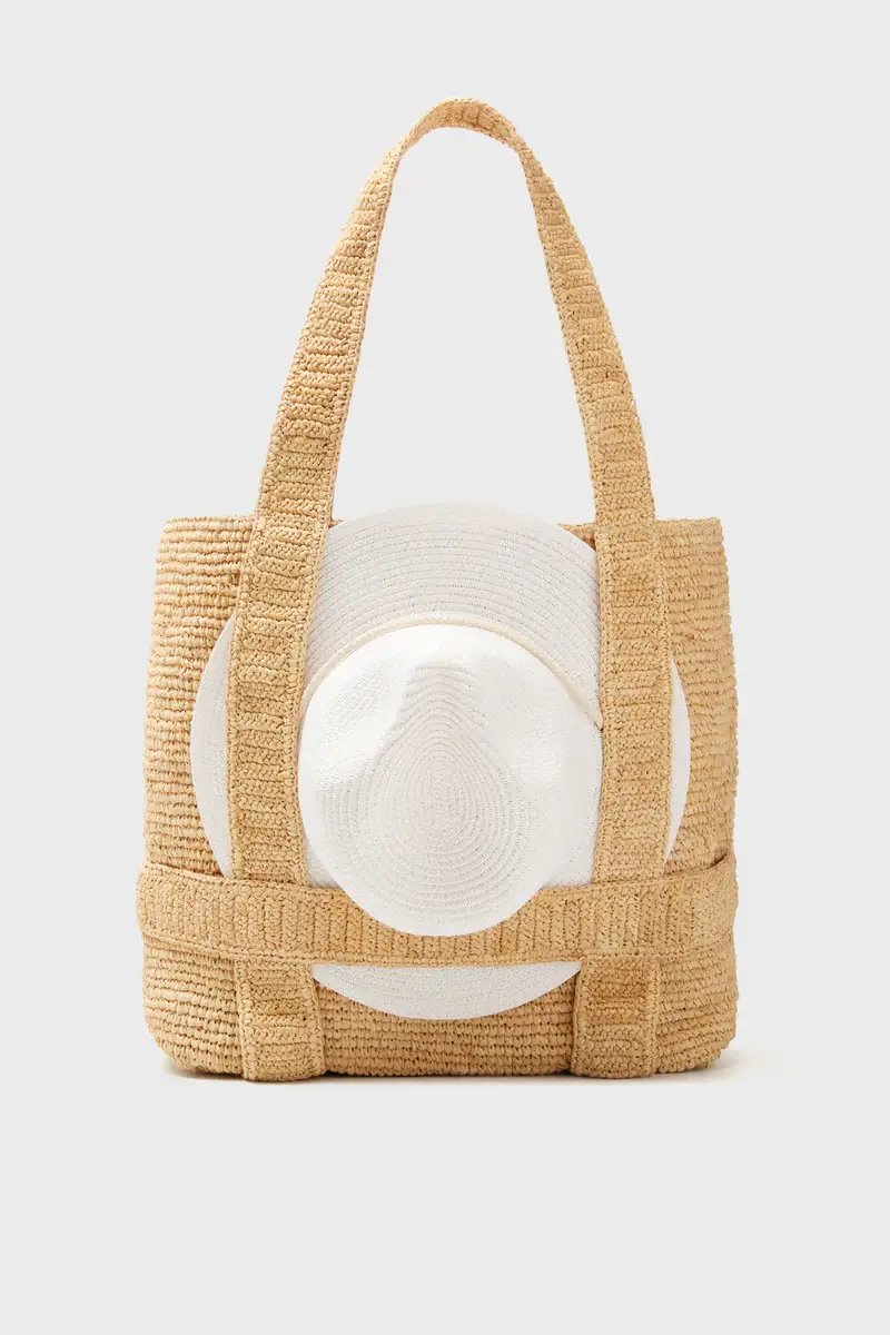A woven straw tote bag with two handles, featuring a white circular sun hat attached to the front—perfect for adding a touch of stylish summer flair to your look.
