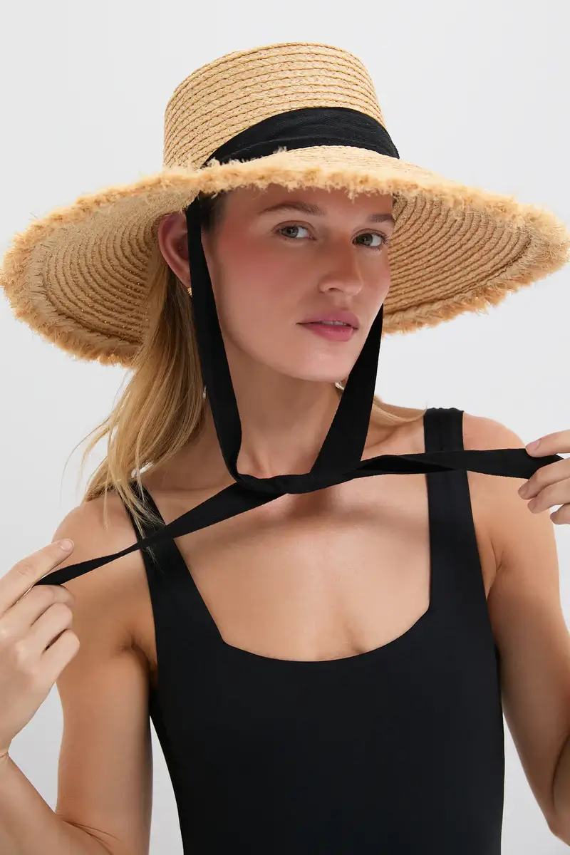 A woman wearing a wide-brim straw hat with a black ribbon ties it under her chin, showcasing stylish summer flair. She is dressed in a sleeveless black top, perfect for chic summer fashion.