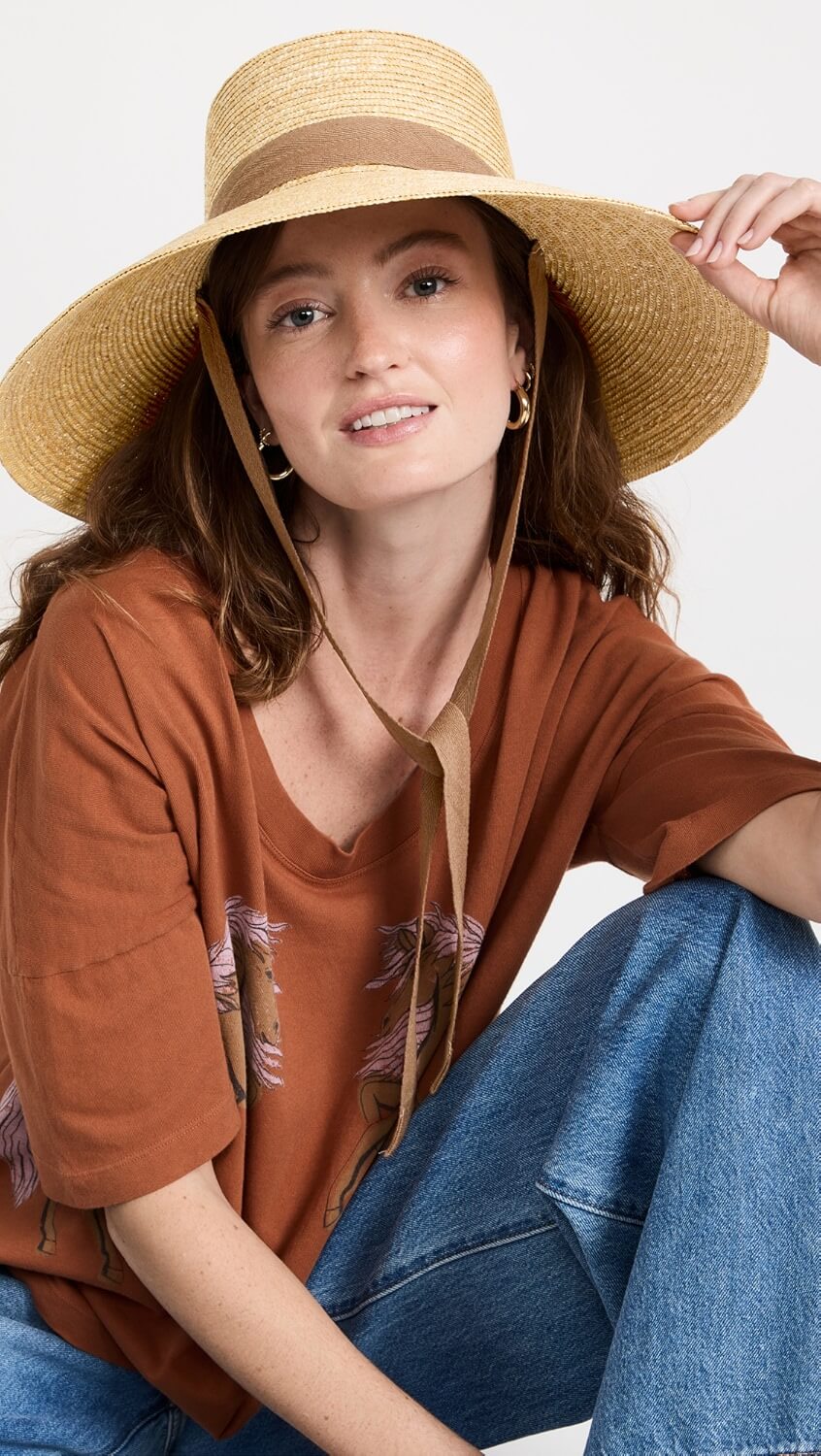 A woman showcases stylish summer fashion in a wide-brim straw hat, a brown t-shirt with a horse design, and blue jeans as she sits and smiles at the camera.