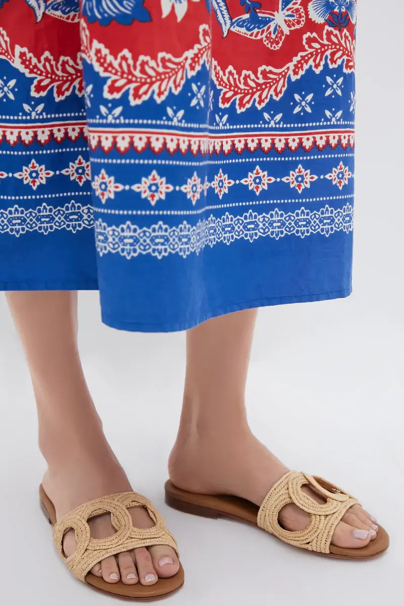 A person wearing a blue and red patterned skirt and beige woven slip-on sandals stands on a light-colored floor, embodying effortless summer style.