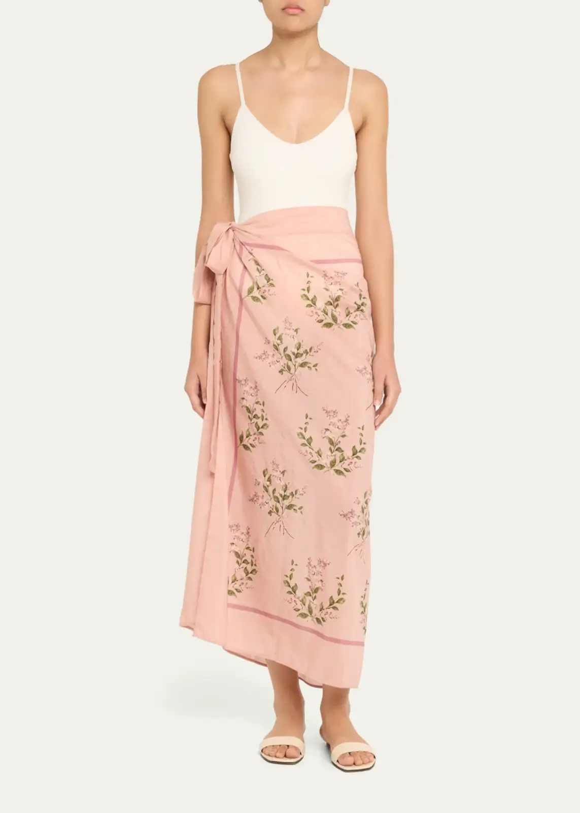 A stylish person wears a white sleeveless bodysuit, a pink floral wrap skirt tied at the waist, and flat sandals, perfect for summer, standing against a plain background.