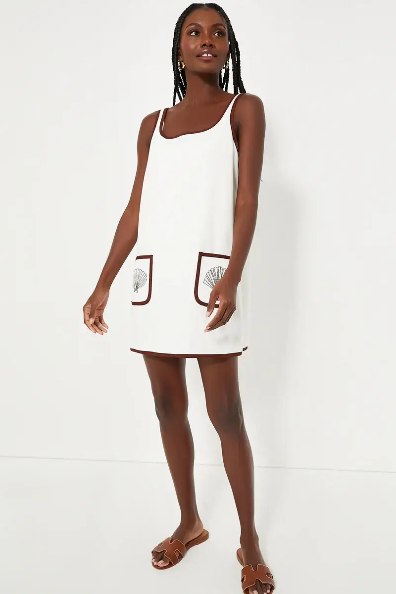 A stylish woman stands against a white background wearing a sleeveless white shift dress with brown trim and two front pockets, paired with brown sandals—a perfect look for summer.