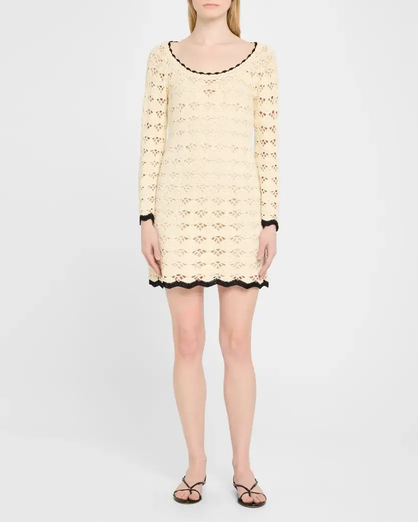 A stylish woman stands facing forward wearing a long-sleeve, cream-colored crochet mini dress with black edging, and black sandals—perfect for a summer day.