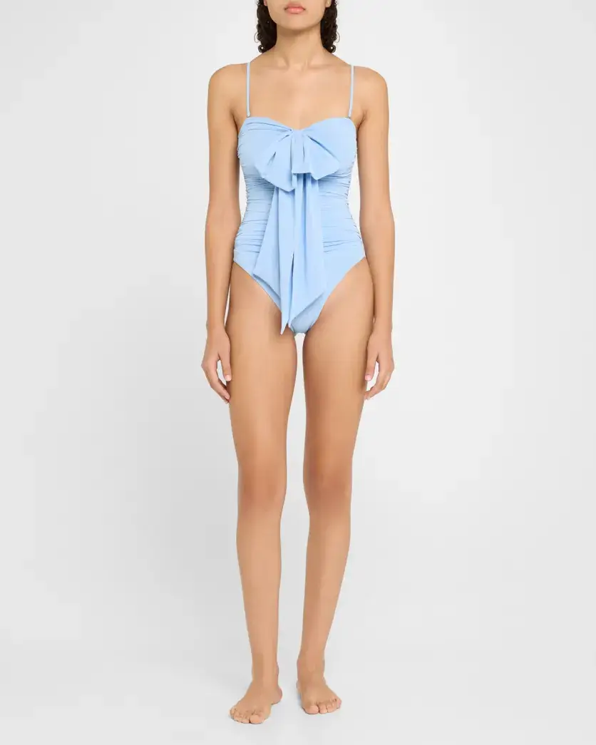 A stylish woman stands barefoot in a light blue one-piece swimsuit with thin straps and a large front bow detail, embracing effortless summer vibes against a plain white background.