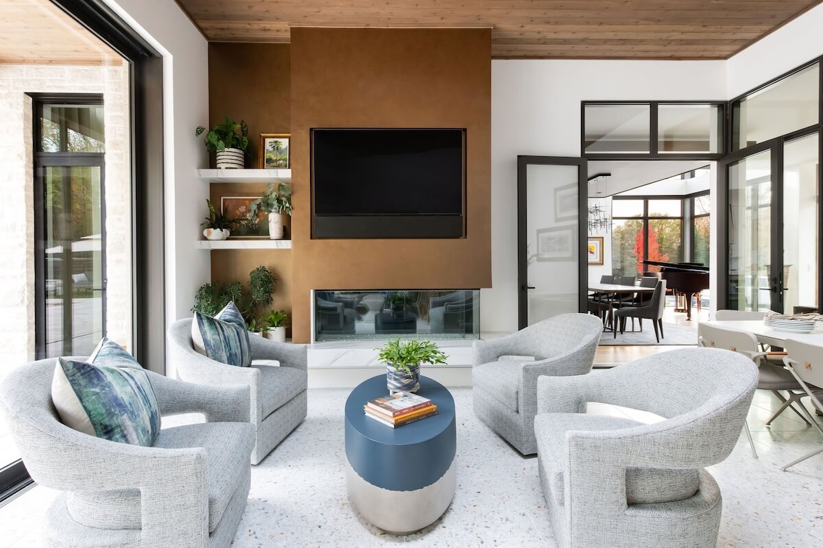2 Homes by Beth Haley Design: A Study in Style & Contrast - 6 This modern living room, curated by Beth Haley Design, features four gray swivel chairs around a round table, a wall-mounted TV above a stylish fireplace, and shelves adorned with plants. Large windows flood the space with natural light.