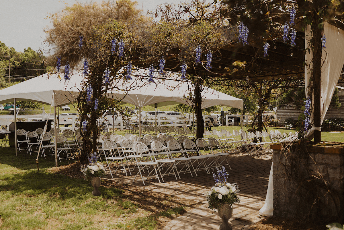 Rows of white folding chairs are arranged under a canopy and pergola adorned with hanging purple flowers at Wildwood Resort, beautifully set up outdoors for an unforgettable event.