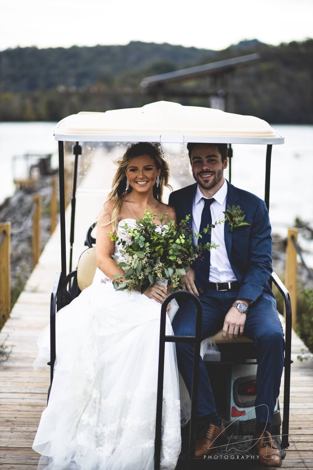 A bride in a white dress and a groom in a suit sit together on a golf cart at Wildwood Resort, smiling, with lush greenery in the background.